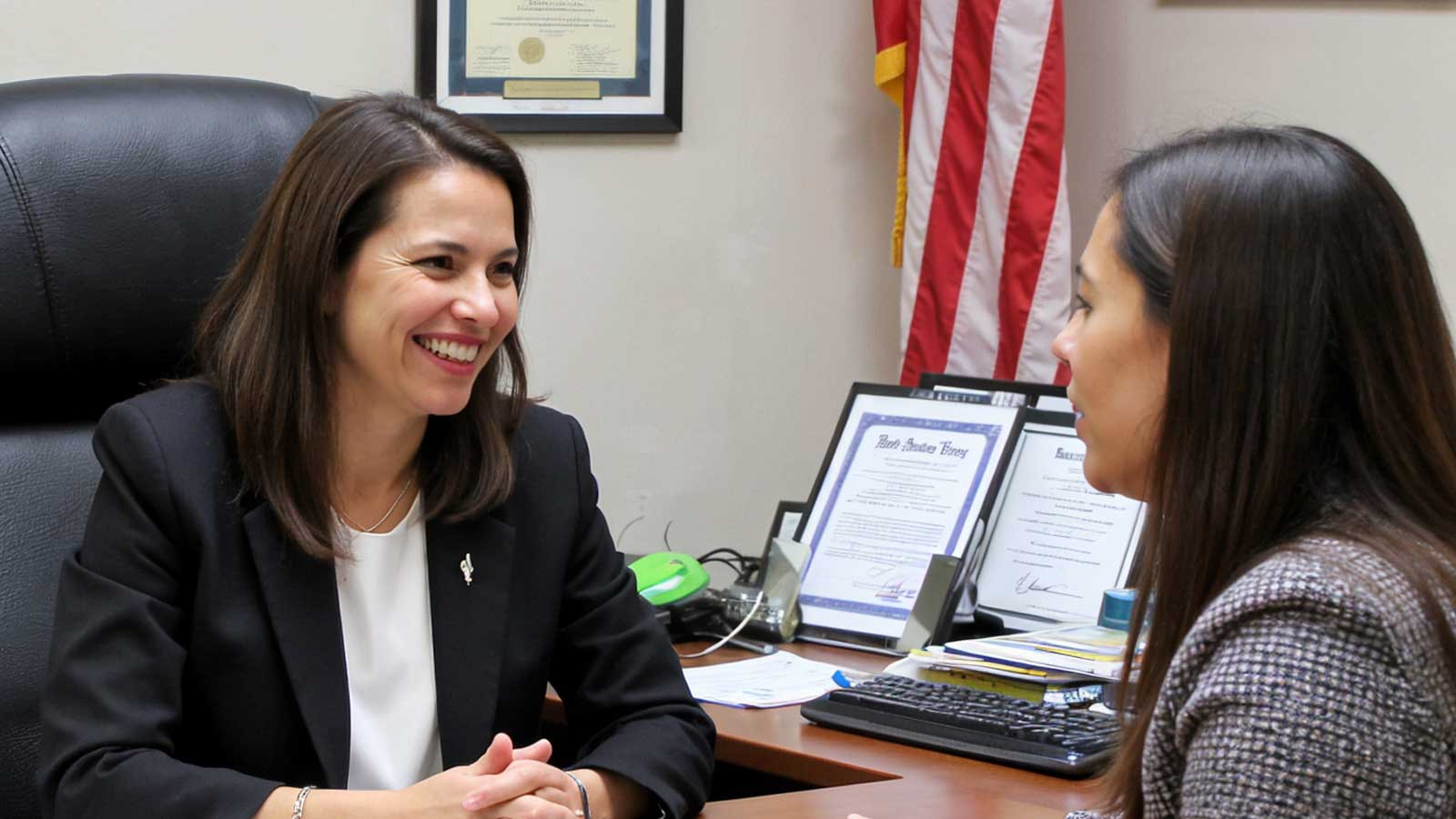 Attorney Karen Monrreal smiles as she talks with a client across her desk, with an American flag and framed certificates.