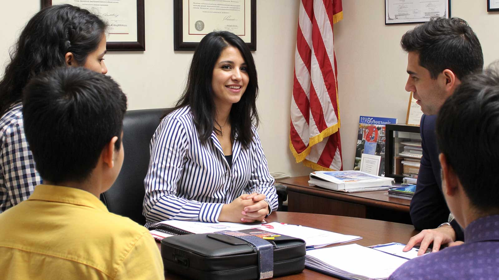 A smiling attorney in a striped blouse meets with a family of four around her desk, with an American flag behind her.