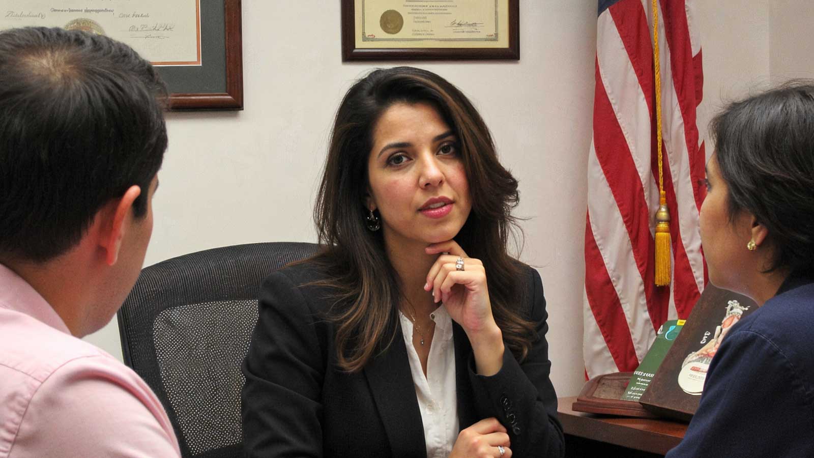 Attorney Karen Monrreal listens closely to two clients at her desk, with framed diplomas and a flag behind her.