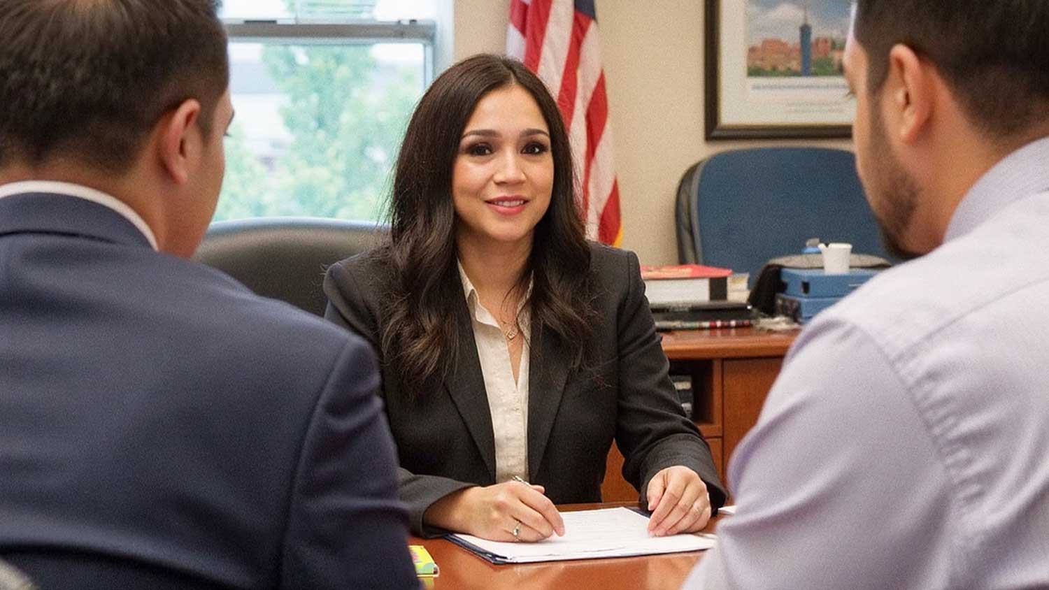 Attorney Karen Monrreal sits at her desk with a pen and papers, meeting with two men across from her.